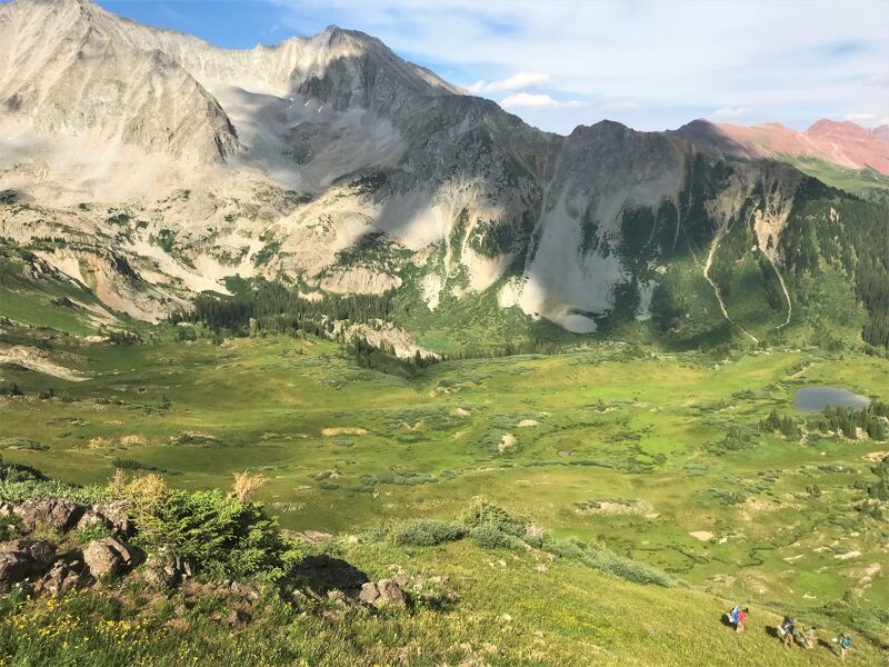 The image shows a scenic mountain landscape with a lush green valley. Towering mountains with rocky peaks and patches of snow dominate the background. The valley is filled with vibrant green grass and scattered trees, with a small lake visible in the distance. In the foreground, there are some rocks and vegetation, and a few small figures of people can be seen hiking on a grassy slope.